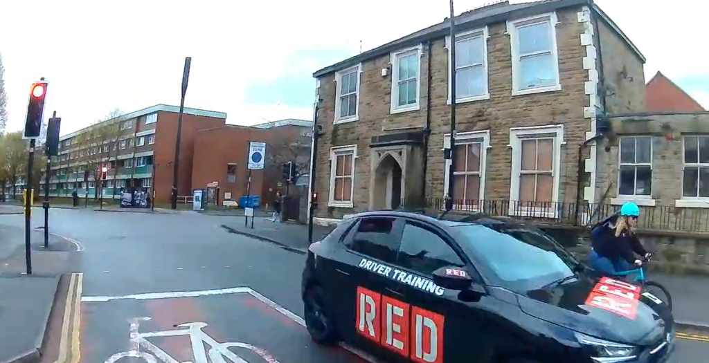 Driver of RED Driving School car races through red light and close passes cyclist