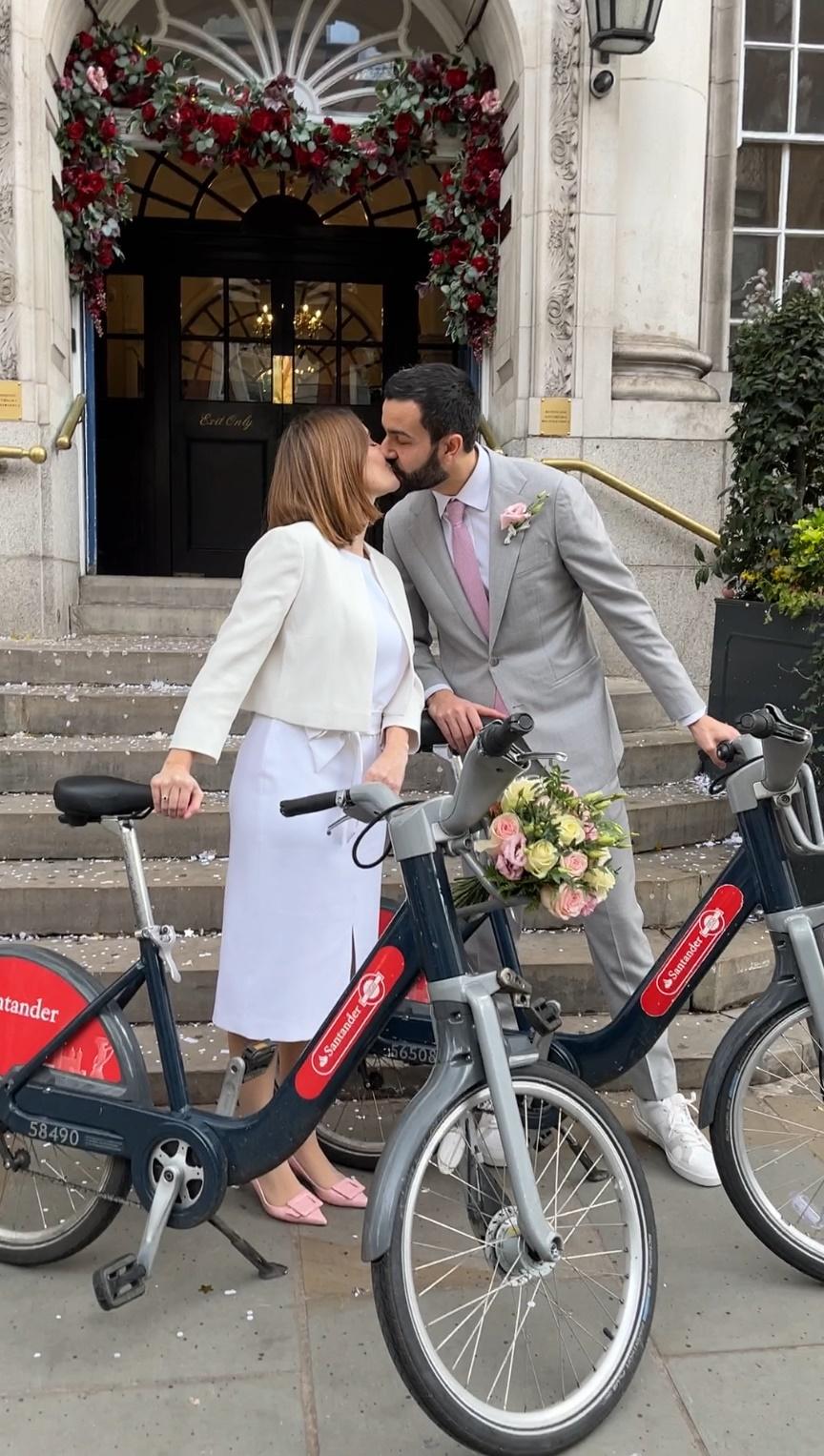 Nicolas Moura and his wife with Santander Cycles on the steps of Chelsea Old Town Hall surrounded by flowers of their wedding day in 2024